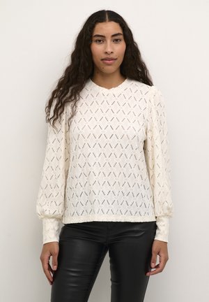 Cream long-sleeve sweater featuring a diamond pattern, textured fabric, and ribbed cuffs, paired with black fitted pants.