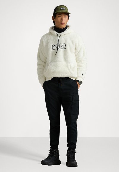 Hvit fleece hettegenser med svart "Polo Ralph Lauren" logo, svarte cargo bukser, grønn caps og svarte støvler. Teksturert, uformell passform.