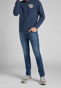 Mörkblå sweatshirt med vit grafisk text, matchad med mörka jeans och grå sneakers med en flerfärgad sula.