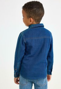 Ragazzo giovane volto di lato che indossa una camicia di denim blu e jeans blu su uno sfondo bianco semplice.