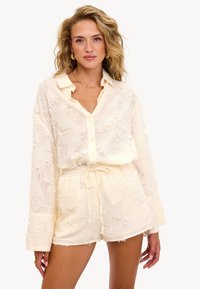 Femme aux cheveux blonds ondulés portant une chemise boutonnée texturée couleur crème et un short à cordon assorti, accessoirisée avec des boucles d'oreilles en or et un collier.