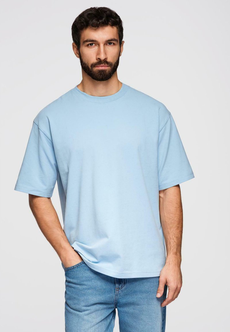 Homme barbu portant un T-shirt ample bleu clair et un jean bleu, debout avec une main dans la poche devant un fond uni.