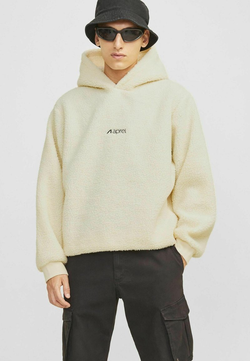 Cremefarvet fleece hoodie med en tekstureret finish, der har et centralt logo. Parret med sorte cargos og en sort bucket hat.