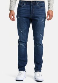 Vaqueros de mezclilla azul oscuro con corte slim, que presentan áreas desgastadas y un desvanecimiento sutil. Combinados con zapatillas blancas para crear contraste.