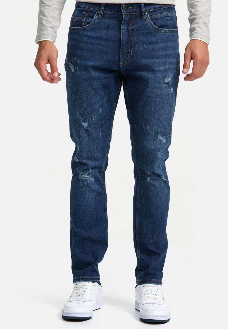 Vaqueros de mezclilla azul oscuro con corte slim, que presentan áreas desgastadas y un desvanecimiento sutil. Combinados con zapatillas blancas para crear contraste.
