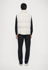 Beige gepolsterte Weste mit hohem Kragen, getragen über einem dunklen Langarmshirt, kombiniert mit dunklen Jeans und weißen Sneakers. Schlichte Designelemente.