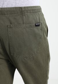 Pantalones de algodón color verde oliva con un bolsillo trasero que lleva la etiqueta "YOUR TURN." Corte recto con costuras visibles y textura suave.