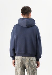 O persoană stă cu spatele la privitor, purtând un hanorac bleu închis și pantaloni cargo cu model de camuflaj, pe un fond alb simplu.