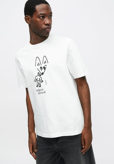 Mladý muž má na sobě bílou tričko Maison Kitsuné s černou abstraktní ilustrací lišky a osoby, v kombinaci s tmavými kalhotami.