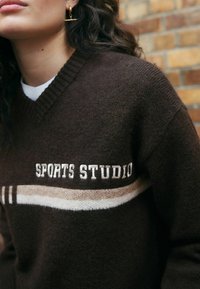 Brązowy wełniany sweter z dekoltem w kształcie v z napisem "SPORTS STUDIO". Posiada kremowo-beżowy paskowany wzór na klatce piersiowej. Miękka tekstura.