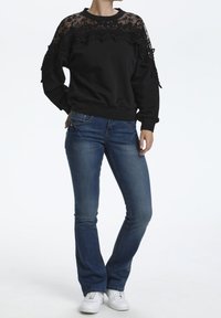Sweatshirt noir avec un empiècement en dentelle florale sur les épaules, associé à un jean flare bleu et des baskets blanches. Tissu lisse et détails texturés.