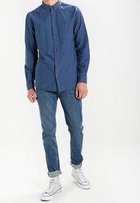 Blaues, langärmliges Hemd mit Knöpfen und einer Brusttasche, kombiniert mit aufgerollten Jeans und weißen Sneakers.