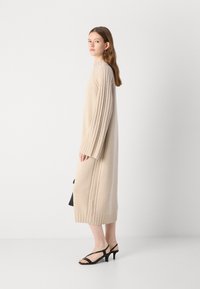 Beige kötött ruha, bordázott ujjakra, oldalszúrásokkal, kerek nyakkivágással és laza fazonnal. Fekete magassarkú szandálokkal párosítva.
