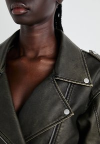 Kurtka ze skóry w oliwkowym zielonym kolorze z srebrnymi zatrzaskami i detalami na suwak, charakteryzująca się klasycznym motocyklowym designem i fakturowanym wykończeniem.