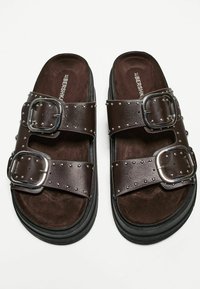 Sandales glissantes brunes avec deux larges sangles, boucles en argent et clous décoratifs. Doublure intérieure douce et semelle noire robuste.