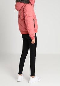 Doudoune rose avec une capuche bordée de fausse fourrure, motif matelassé diagonal, poche zippée à l'avant et poignets côtelés. Associée à un legging noir et des baskets blanches.