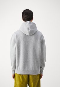 Helle graue Kapuzenpullover mit weicher Textur, gerippten Bündchen und Saum sowie lockerer Passform, getragen über senfgelben Hosen.