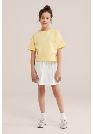 Jong meisje staat met ontspannen handen, draagt een geel T-shirt met korte mouwen en bloemdetails, een witte rok, witte sokken en zilveren schoenen.