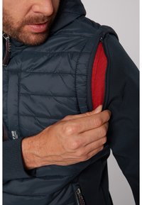 Vesta puffer albastră închis cu căptușeală roșie, dotată cu buzunare cu fermoar și glugă, cu un design texturat și croiala elastică a unei guri de mână.