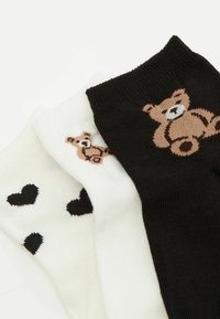 Trzy pary skarpetek kostkowych: jedna czarna z grafiką misia, jedna biała z grafiką misia oraz jedna biała w czarne wzory serc.