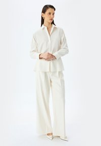 Camisa blanca con un patrón texturizado, cuello abotonado y mangas largas, combinada con pantalones de pierna ancha en un color a juego y zapatos de punta.