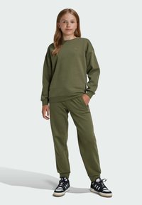 Maslinasto zeleni dukserica i odgovarajuće hlače od mekog materijala, s okruglim izrezom, rebrastim manžetama i suptilnim logotipom na grudima. Crno-bijele tenisice.