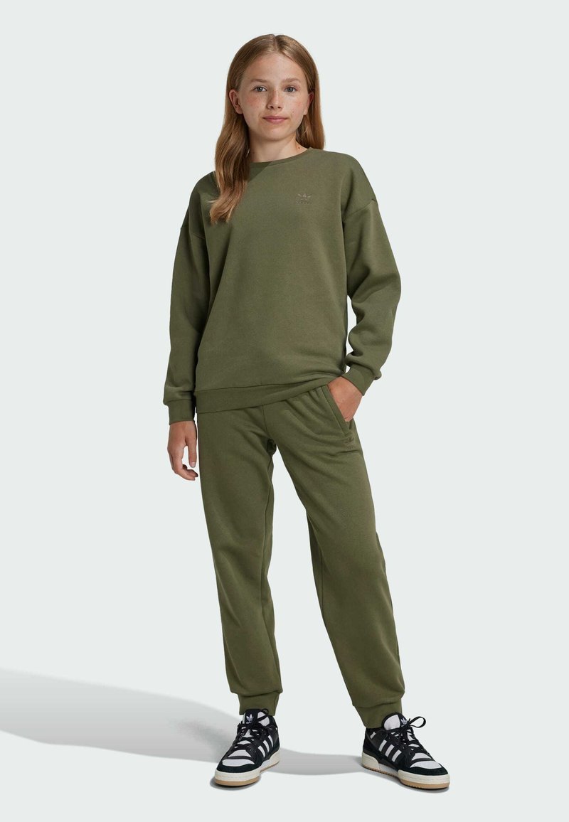 Maslinasto zeleni dukserica i odgovarajuće hlače od mekog materijala, s okruglim izrezom, rebrastim manžetama i suptilnim logotipom na grudima. Crno-bijele tenisice.