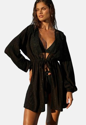 Woman with wet hair wearing a black, long-sleeve, sheer cover-up with floral lace over a black bikini, standing with one hand on her hip.