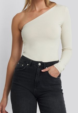 Frau trägt ein cremefarbenes, einseitig schulterfreies Langarm-Top kombiniert mit hochtaillierter schwarzer Jeans, Hand in der Tasche.