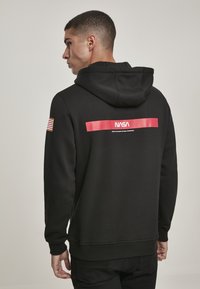 Sweatshirt à capuche noir avec logo et texte NASA rouges au dos, présentant un patch du drapeau américain sur la manche gauche. Tissu doux, coupe décontractée.