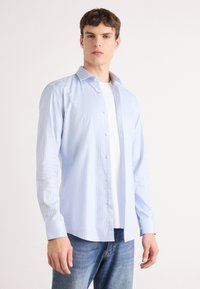 Camisa de botón azul claro con rayas blancas verticales, de mangas largas y cuello extendido, sobre una camiseta blanca, combinada con jeans azules.
