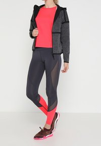 Tenue de sport comprenant un sweat à capuche noir zippé, un haut ajusté rose vif et un legging noir avec des accents roses et en maille. Baskets marron avec des semelles roses.