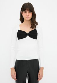Weißes Langarmoberteil mit einer breiten schwarzen Schleife am Ausschnitt, glatte Textur, taillierter Schnitt, kombiniert mit schwarzen hochgeschnittenen Hosen.