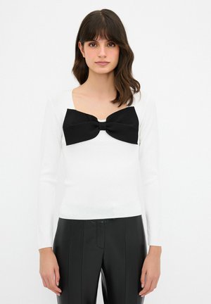 Maglietta bianca a maniche lunghe con un ampio fiocco nero sul colletto, texture liscia, design aderente, abbinata a pantaloni neri a vita alta.
