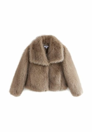 Personne portant une veste en fausse fourrure marron clair sur un haut rouge et un pantalon noir, debout à l'intérieur devant un mur neutre.