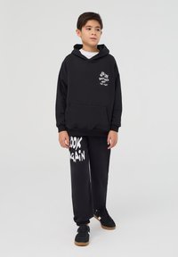 Felpa nera con tasca frontale e testo grafico bianco. Abbinata a pantaloni della tuta neri con "GUARDA ANCORA" in grande carattere bianco. Sneakers nere.