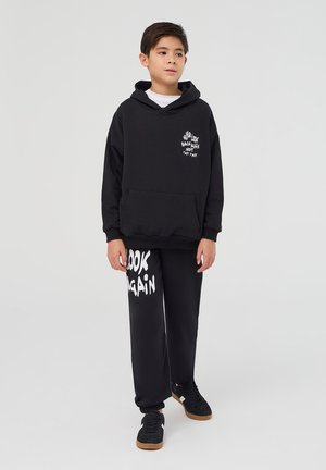 Felpa nera con tasca frontale e testo grafico bianco. Abbinata a pantaloni della tuta neri con "GUARDA ANCORA" in grande carattere bianco. Sneakers nere.