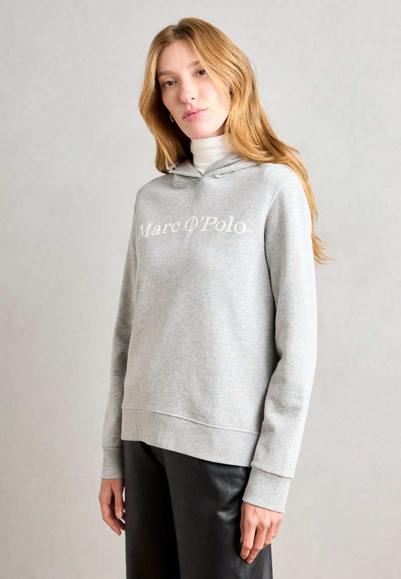 Grauer Kapuzenpullover aus weichem Stoff, mit gerippten Bündchen und einem weißen Logo-Aufdruck "Marc O'Polo" auf der Brust. Kombiniert mit dunklen Hosen.