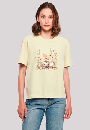Light yellow cotton t-shirt featuring a printed design of a duckling among floral elements and decorated eggs, with short sleeves and a relaxed fit.