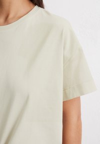Jasno beżowy t-shirt z okrągłym dekoltem i krótkimi rękawami. Wykonany z miękkiego, gładkiego materiału, charakteryzujący się luźnym krojem i minimalistycznym stylem.