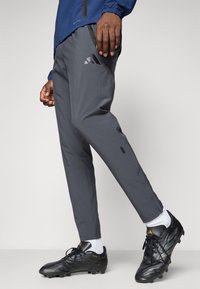 Grå atletiske bukser lavet af letvægtsstof, med en slim fit, sidelommer og et Adidas-logo på venstre lår, parret med sorte fodboldstøvler.