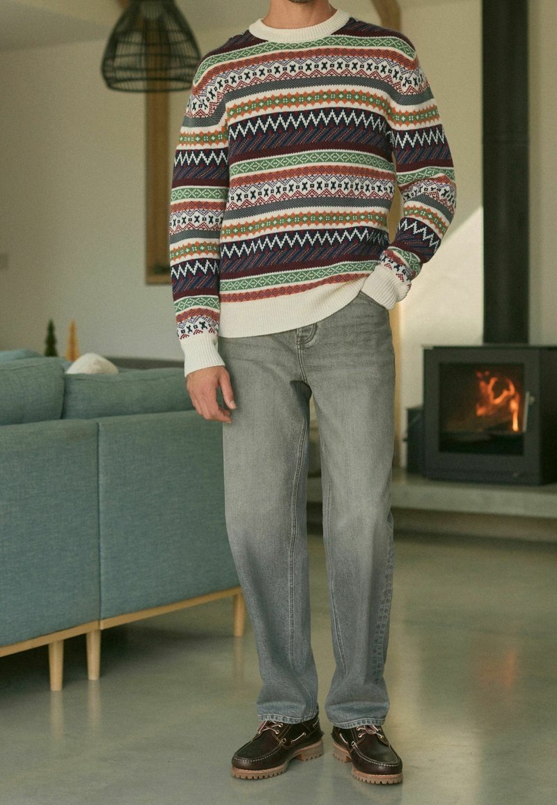 Homme portant un pull à motifs, un jean gris et des chaussures marron, debout à l'intérieur près d'une cheminée allumée et d'un canapé vert.