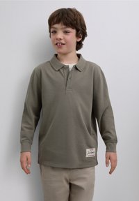 Tricou polo verde măsliniu cu mâneci lungi, cu guler, doi nasturi și detaliu cu etichetă laterală. Textură netedă și croială dreaptă.