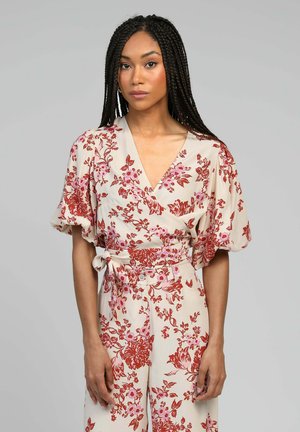 Salopette fleurie avec un décolleté en V croisé, des manches courtes et un fond crème. Présente des motifs floraux rouges et roses avec une taille à nouer.