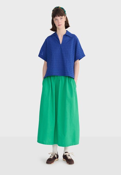 Kobieta o krótkich brązowych włosach nosi niebieską koszulę w kratę z krótkim rękawem, zielone szerokie spodnie, białe skarpetki, brązowe trampki oraz kwiatową opaskę na głowę.