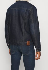 Uomo che indossa un giubbotto di jeans blu scuro con colletto in shearling e jeans blu scuro abbinati, visto di spalle su uno sfondo neutro.