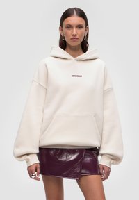 Felpa oversized color crema con cappuccio regolabile, tasca frontale e logo "WRSTBHVR". Abbinata a una mini gonna lucida color bordeaux. Tessuto liscio.