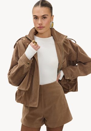 Femme portant une veste en suède marron et un short assorti, un haut blanc à manches longues, une grande boucle d'oreille et des bagues en or, posant avec la main près du col.