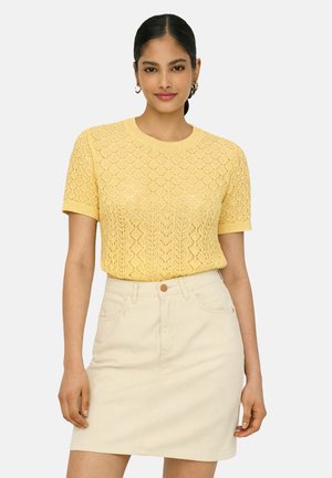 Mujer con un top de punto amarillo de manga corta con un patrón de diamantes y una falda beige de cintura alta, de pie contra un fondo liso.