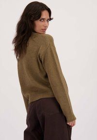 Olijfgroene gebreide trui met een ronde halslijn, ribgebreide manchetten en zoom, en een comfortabele pasvorm. Gecombineerd met donkerbruine broeken.